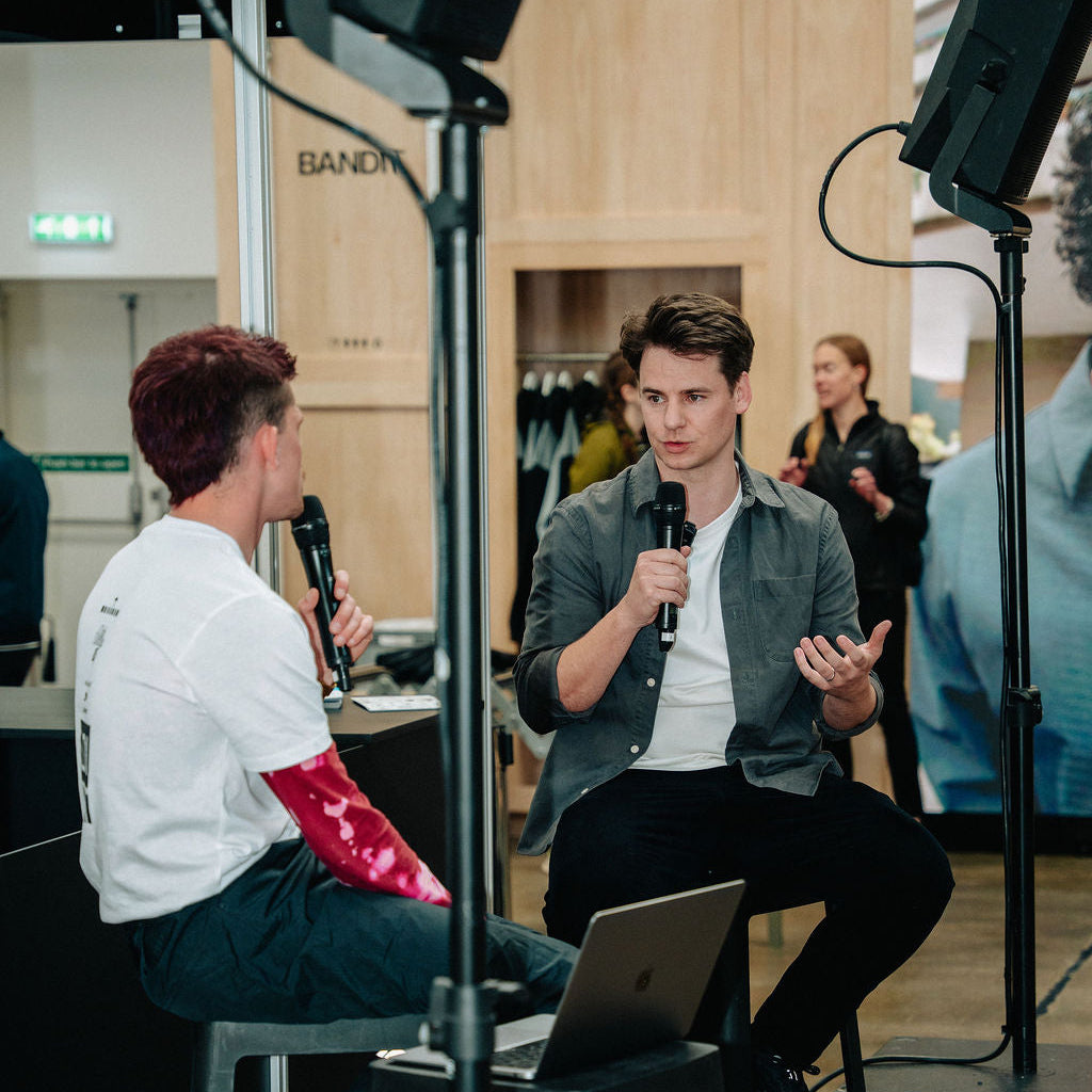 Two people sitting on stools with microphones and a laptop in a room with wooden walls.